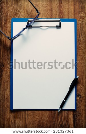 Eye-glasses with black frames and a black pen on an empty white page clipped in a blue clip folder, on a wooden table, high-angle close-up