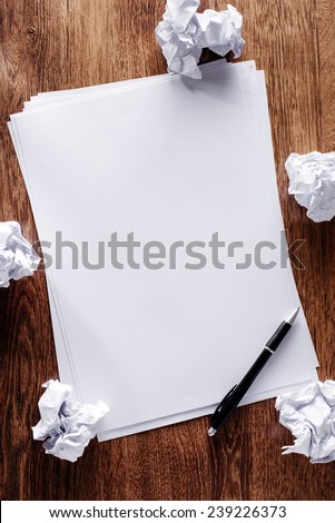 Overhead view of a stack of blank white sheets of paper surrounded with crumpled remnants screwed up in frustration on a wooden desk