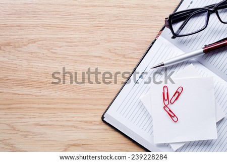 Glasses, pen, clips, notebook lie on light brown desktop