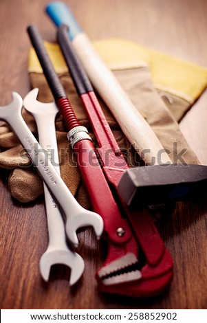 Close up Hand Working Tools like Open Ended and Pipe Wrenches, Cross Pein Sledge Hammer and a Pair of Gloves on Top of a Wooden Table.