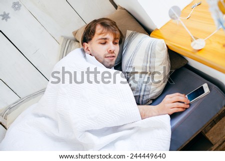 Sleepy young man in bed extending hand to alarm clock