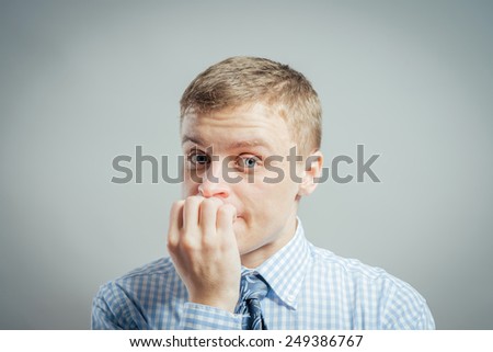 Closeup portrait of young man in blue shirt biting his nails and looking at you with a craving for something or anxious, isolated on  gray background. Negative Human facial expressions