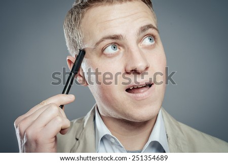 Thinking man with pen near his face isolated on gray background. Closeup portrait of a casual young pensive businessman looking up at copyspace.
