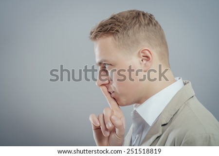 Closeup portrait young serious businessman placing finger on lips saying, shhh, be quiet, silence, isolated grey background. Facial expression, human emotions, sign, symbol, body language, perception