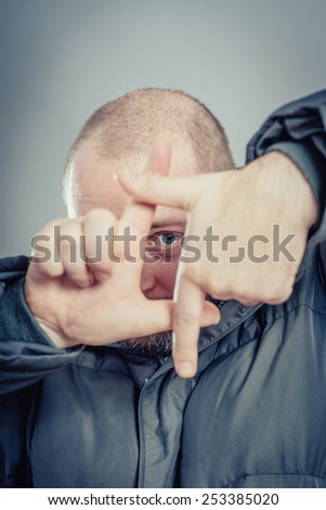 Focusing at you. Playful young man in  gesturing finger frame and smiling while standing against grey background