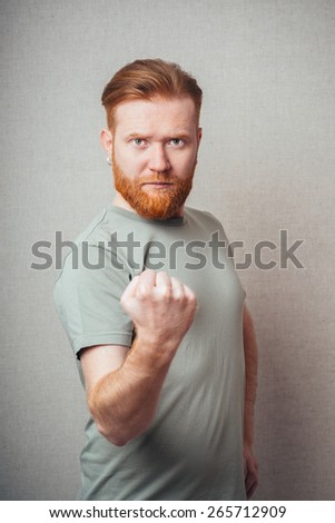 Unleashing his emotions.  Portrait of handsome Young hipster red bearded man wearing orange tshirt and showing his fist with angry face expression while standing against white background