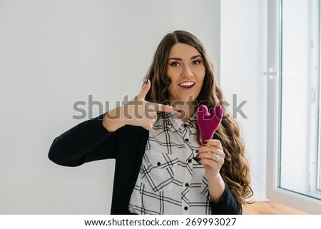 Picture of a small pink heart in hands, lonng hear brunette female holds handmade heart soft toy, woman with Valentine gift against the window, happy girl smiling