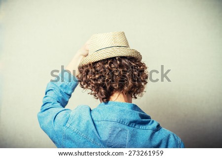 Curly young man in a wicker hat over his head put his hands resting. Back view. On a gray background