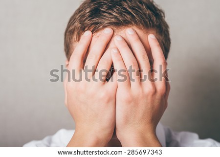 Shocked and terrified. Portrait of young man covering his face by hand and looking at camera while standing against grey background