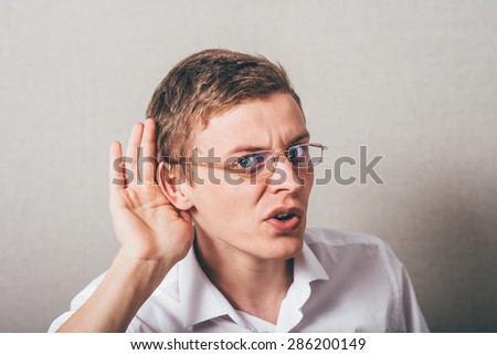 The man in glasses shows a gesture louder, I can not hear, hand near ear. On a gray background.