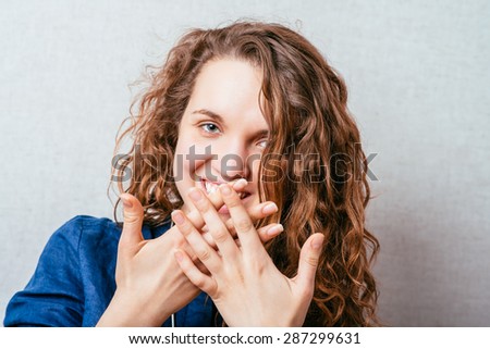 Curly smiling woman covers her mouth with her hands. Gray background.