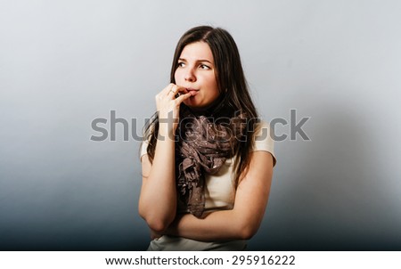 Young woman is thinking and nervous hand to her mouth. On a gray background.