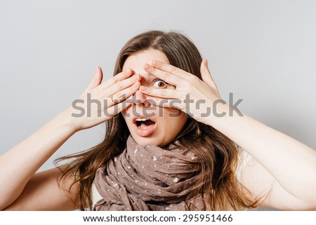 A young woman scared, looking through her fingers. On a gray background.