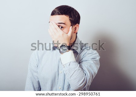 Shocked and terrified. Portrait of young man covering his face by hand and looking at camera while standing against grey background