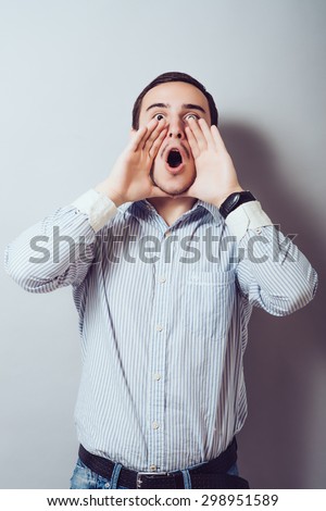 Great news! Cheerful young man in holding hands near mouth and shouting while standing against a gray background