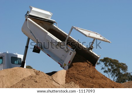 A white dump truck full of dirt lifts the back end and lets the soil go.