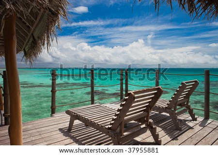 Two wooden lounge chairs sitting on the deck of an overwater bungalow suite overlooking the lagoon of a tropical paradise in Tikehau, Tahiti.