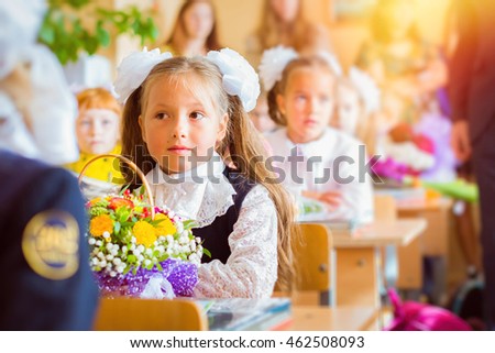 Little first-grader, girl-student goes to school on knowledge day - September First. Student of elementary school in uniform with bows posing in classroom for student Desk, bag with textbooks. Russia