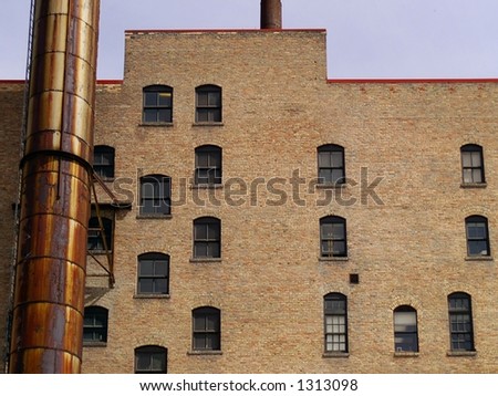 Windows and a rusted smokestack against a brown or tan brick wall on the side of an urban apartment building.