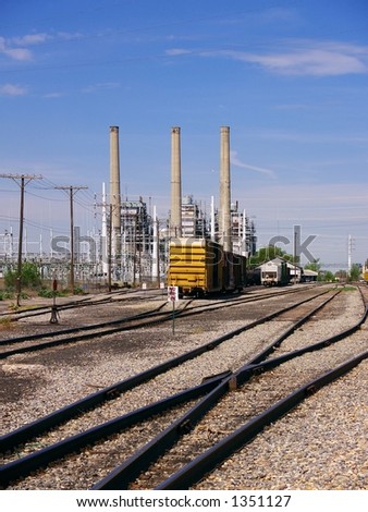 Rail lines run across a desolate industrial area with smokestacks and power lines in the background.