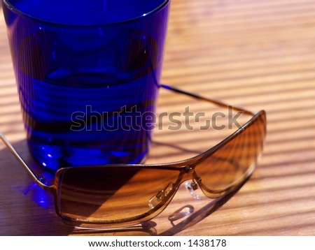 Womens\' sunglasses resting next to a dark blue cocktail tumbler in strong light in shallow focus.