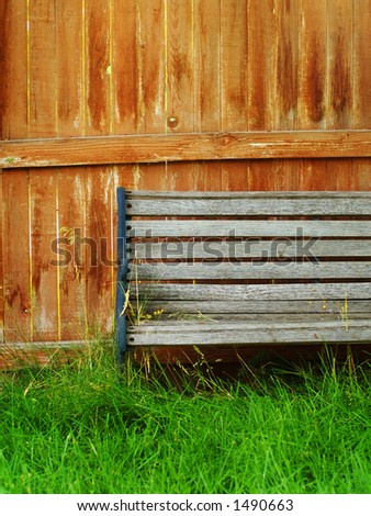 Faded wooden bench in front of bright, worn wooden fence amidst bright green grass.