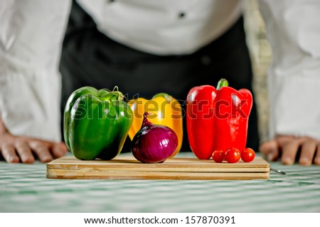 chef  stand in front of a table with apron and white tunic making a salad with colorful vegetables