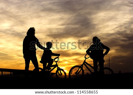 A brother and sister stand with their bikes along with their mother while watching the sunset.