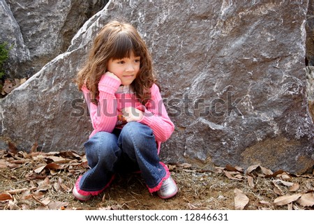 A young girls sits in a squat infront of a large rock.