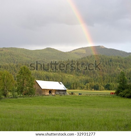 A rainbow falls over a barn in a farm field.