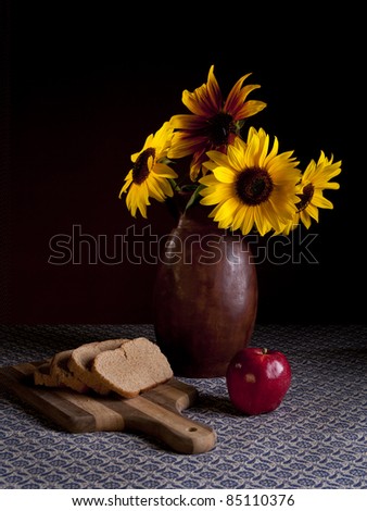 A fine art image of a vase of sunflowers, bread, and an apple.
