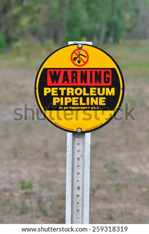A red, black and yellow sign on a post reading Warning Petrolium Pipeline against a natural background