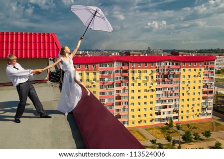 The newly married couple with white umbrella standing on the roof of house under cloudy sky