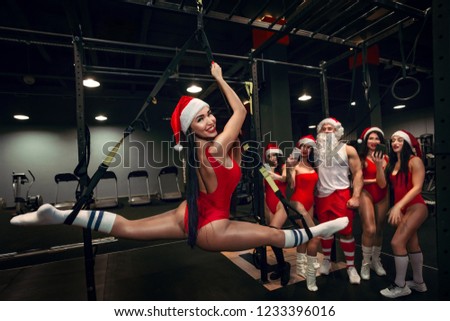 Young attractive Santa woman does splits crossfit stretching with trx fitness straps in the gym\'s studio and Santa with helper making selfie in Gym