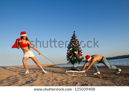 Sexy Santa helper  pulling Santa on a sled with Christmas tree at the beach.  (concept: Tropical winter fun)