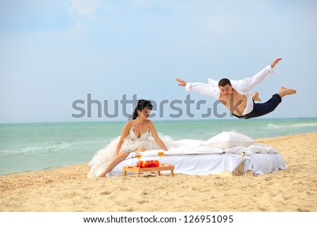 Happy groom flying on bed to his sweetheart on the beach