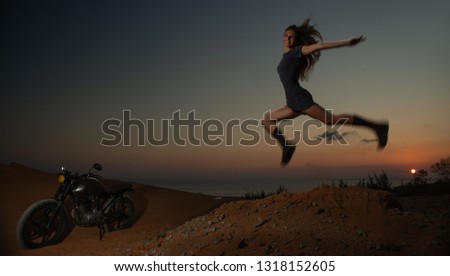 Woman jumping for joy next to motobike at sunset.