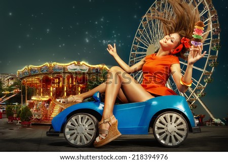 Pretty woman driving toy car holding ice cream and smile.  Amusement park at night - ferris wheel  in motion