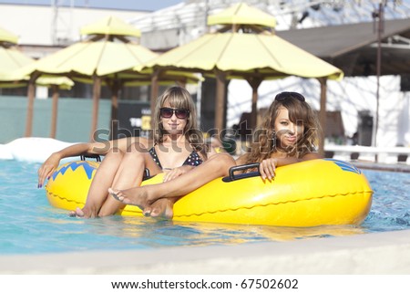 Two attractive slim and tanned young ladies lying on inflatable ring on sunny swimming pool on vacation or holiday