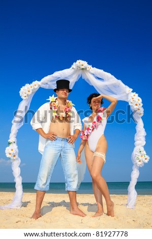 Groom with bride wearing lei, standing under archway on beach, peering into the distance