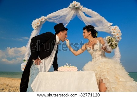 Bride and Groom Under Archway on Beach with wedding cake. Bride giving a bit of cake to groom