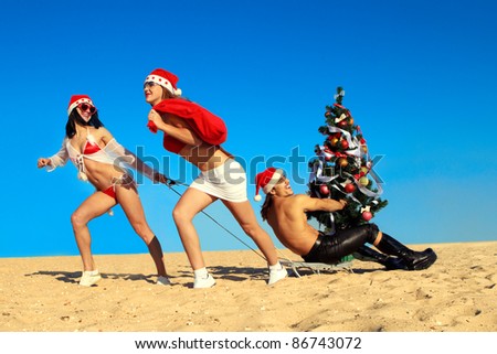 Two Sexy Santos  pulling Santa on a sled with Christmas tree at the beach.  (concept: Tropical winter fun)