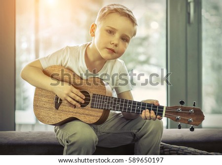 Young boy play on guitar at home at sunny day. Boy play on ukulele - hawaiian guitar.