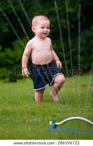 Toddler cools off by playing in water sprinkler at home in his back yard on hot summer day