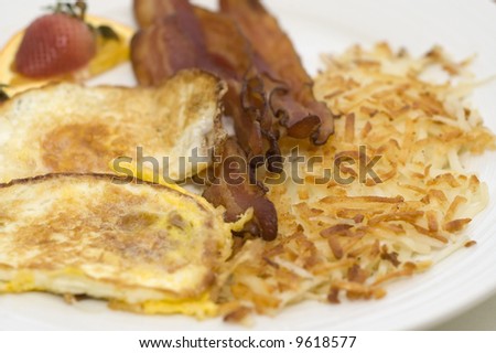 Bacon and fried egg breakfast with hash browns and fruit.  Shallow depth of field with focus on the bacon.