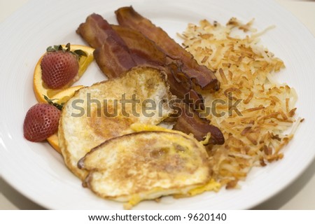 Bacon and fried egg breakfast with hash browns and fruit.  Shallow depth of field.