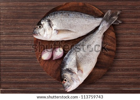 Two fresh fish on round wooden chopping board on brown background, top view. Culinary seafood concept in natural brown.