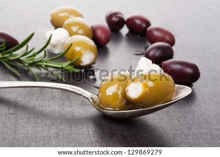Cheese filled green and black olives, antique silver spoon, feta cheese pieces and rosemary herbs with extra virgin olive oil on dark grey and black background. Luxurious culinary appetizer concept.