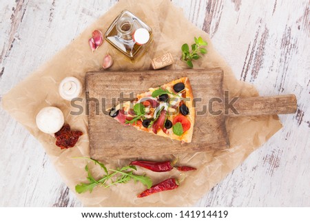 Culinary pizza cooking. Pizza piece on wooden kitchen board, with various ingredients and toppings. Fresh herbs, olive oil, garlic, edible mushrooms, dried tomatoes and chili pepper. Pizza eating.