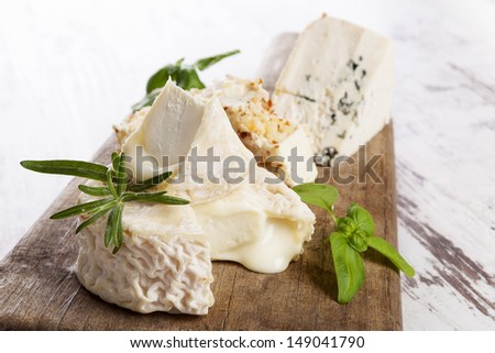 Various goat cheese and blue cheese with fresh herbs on wooden kitchen board on white wooden background.